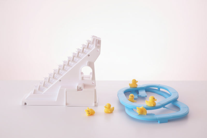 Toy track with a white and pink design on a wooden surface, next to a clear plastic package with a blue toy track and yellow ducks.