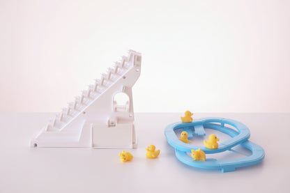 Toy track with a white and pink design on a wooden surface, next to a clear plastic package with a blue toy track and yellow ducks.