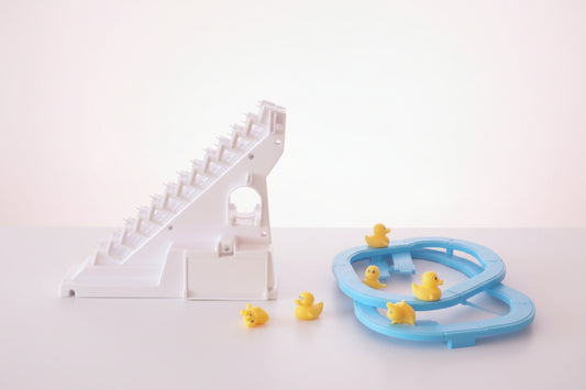 Toy track with a white and pink design on a wooden surface, next to a clear plastic package with a blue toy track and yellow ducks.