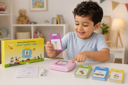 A young boy in a light blue t-shirt is happily interacting with a children's tablet learning device on a white table. He is holding a purple card, with several colorful cards and a yellow educational box displayed around him.