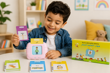 A young boy wearing a denim shirt sits at a white table, smiling as he plays with a pink early education toy device. He holds up a purple card, and several colorful instruction cards and a green-and-yellow product box are spread out in front of him on a soft, warm indoor background.
