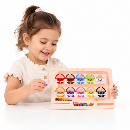 A young girl with brown hair in a ponytail smiles while playing with a pink plastic bead-sorting toy. The toy has multiple colorful baskets with cartoon faces, and she is holding a purple bead.