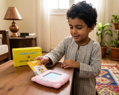 A young boy with curly dark hair sits at a wooden table in a warmly lit room, interacting with a pink handheld learning device and a yellow Arabic alphabet card. A yellow box labeled with Arabic text is visible behind him.