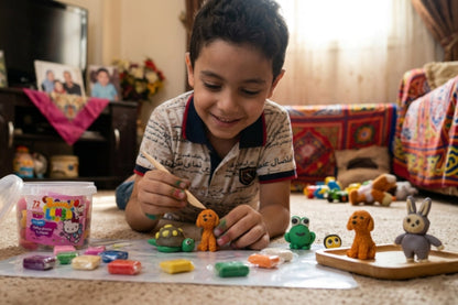 A young boy sits on a carpeted floor in a living room, painting small clay figurines with a wooden stick. A tub of colorful clay modeling compounds and several painted figurines are spread out on a plastic sheet in front of him.