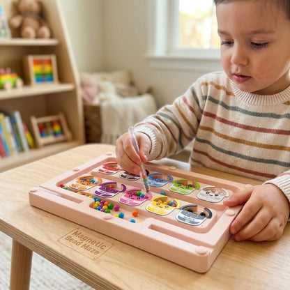 A young child wearing a striped sweater sits at a wooden table, playing with a pink Magnetic Bead Maze toy. The child is holding a clear pen-like tool over a tray filled with colorful magnetic beads.